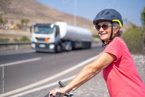 Portrait of happy mature woman, wearing protective helmet, on her bike in the sunny road. Healthy and active retirement concept