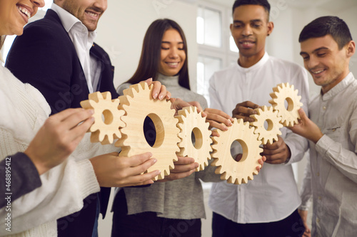 Team of happy young diverse people joining gears in chain as metaphor for good, well-coordinated group work, effective business system, teamwork, interaction, participation, involvement, collaboration