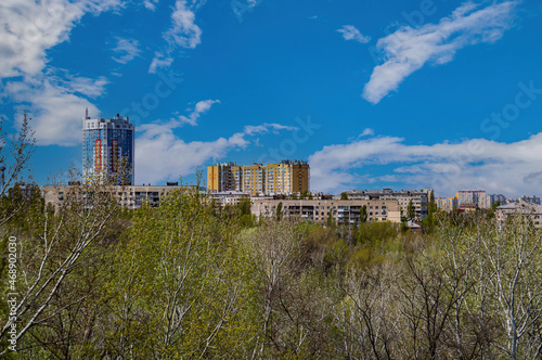 Urban residential buildings on the horizon from above. City landscape. Blue sky with white clouds. The tops of the trees. Bird s-eye view. Cloudy horizon. Background image.