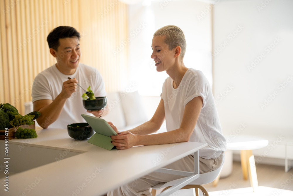 Caucasian woman watching something on digital tablet while her boyfriend eating salad. Concept of healthy and vegetarian eating. Domestic lifestyle. Smiling multiracial couple in studio apartment