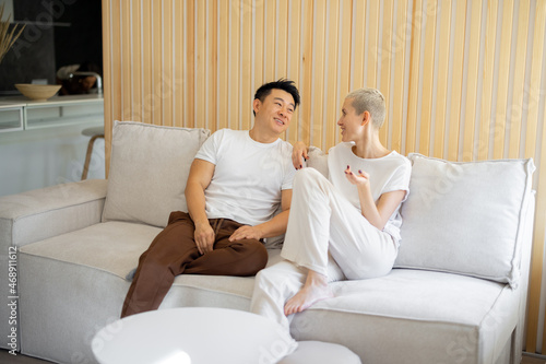 Asian man and caucasian woman sitting and talking on sofa at home. Concept of relationship and spending time together. Idea of domestic lifestyle. Smiling multiracial couple in studio apartment