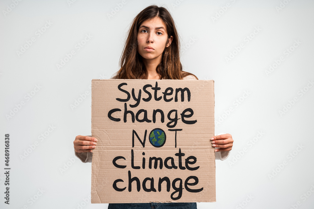 Brunette worried woman pointing at the carton placard with System change not climate change writing. Concept of nature destruction and eco activism Brunette worried woman pointing at the carton placard with System change not climate change writing. Concept of nature destruction and eco activism