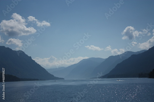 Teletskoye Lake in the Altai Mountains. The water glistens in the sun. Trees grow on the mountain. Blue sky and white clouds