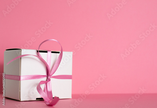 A small gift with a pink ribbon on pink background. White gifts with a pink bow. Christmas gifts. Gift for woman. Holiday pink background with gift, white satin bow, ribbon. Space for text