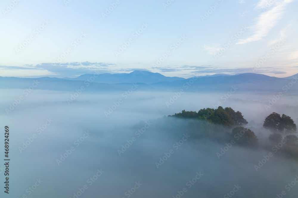 View from above, stunning aerial view of a forest surrounded by fog with a mountain range in the distance.