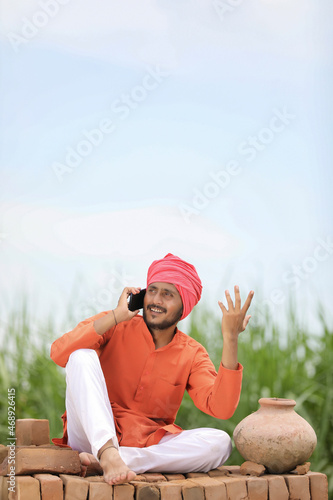 Indian farmer sitting on bricks and talking on smartphone at green corn field.