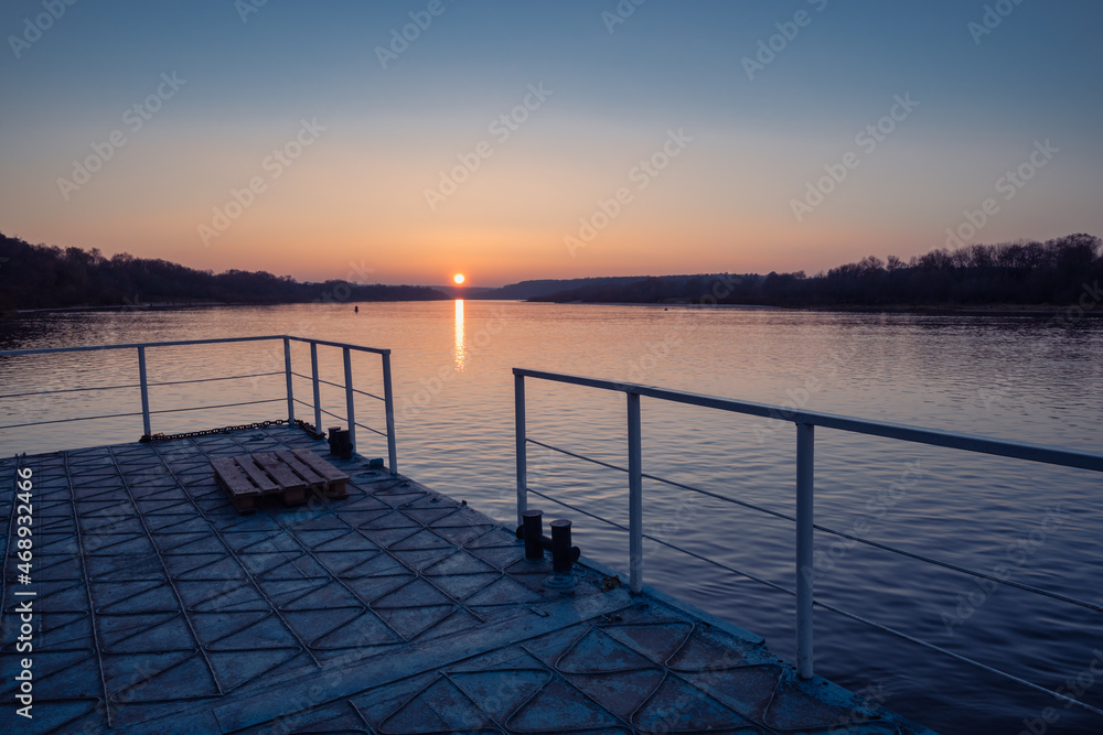 Sunset on a wide river with bright red reflections and a sun disc over the horizon from a metal pier with railing. The rural scene photo from a low point view.