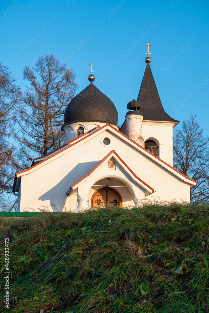 A white stone orthodox church on a hill against clear blue sky and bare trees behind.