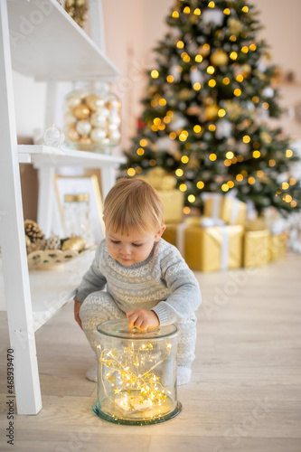 Adorable baby girl sitting near Christmas tree with festive lights and xmas gifts. Chrismas and New Year.