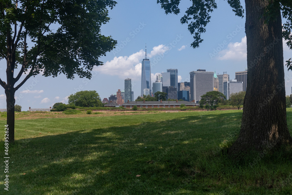 Lower Manhattan Skyline framed between Trees seen from a Green Grass Field on Governors Island in New York City during the Summer Lower Manhattan Skyline framed between Trees seen from a Green Grass Field on Governors Island in New York City during the Summer