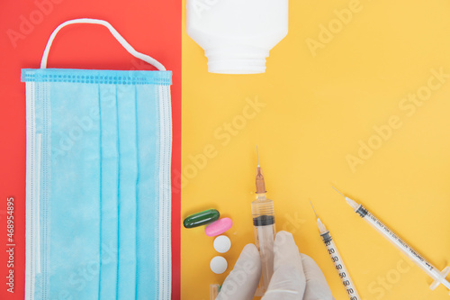 light blue mask on red background, near white pills and capsule beside syringe.