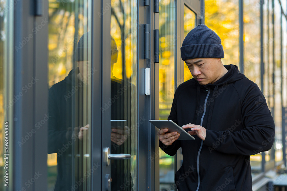 Male thief hacker tries to break the alarm in the house with a virus program, uses a tablet, Asian near the house Male thief hacker tries to break the alarm in the house with a virus program, uses a tablet, Asian near the house
