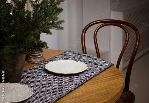 White plates on the tablecloth, fir branches in a vase on a wooden table. A wooden chair stands next to the table. Christmas interior. Table setting. Interior design. Home decoration. New Year concept