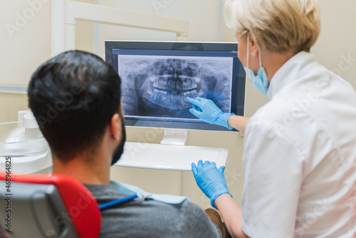 Female dentist orthodontist showing explaining an x-ray photo of jaws on computer screen to a male patient in dental clinic. Stomatology and healthcare concept