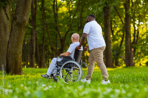 Caregiver and old man in a wheelchair. Professional nurse and patient walking outdoor in the park at sunset. Assistance, rehabilitation and health care. © Acronym