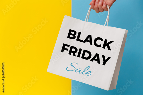 Female hand holding white shopping paper bag with Black Friday Sale text on yellow and blue background. Sale, discount, shopping concept.
