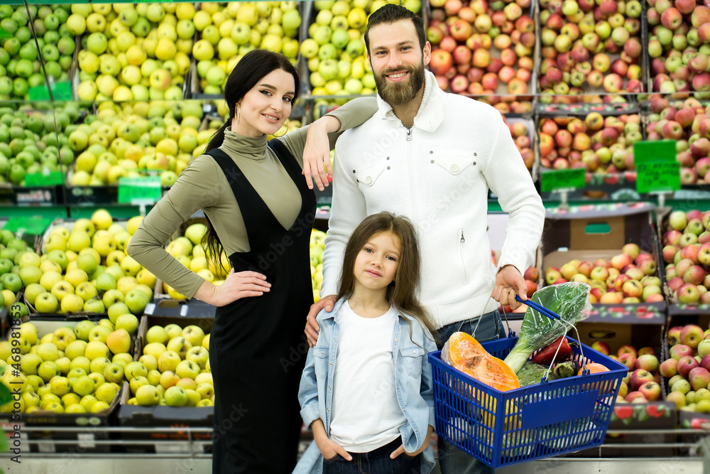 Portrait of a cheerful family standing with a full cart in the supermarket in the vegetable Department on the background of the counter with apples. Portrait of a cheerful family standing with a full cart in the supermarket in the vegetable Department on the background of the counter with apples.