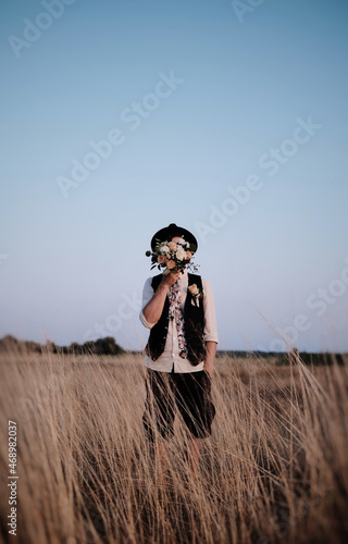 Wedding ceremony in rustic style, man in hat stay in a field and cover his face with flowers.