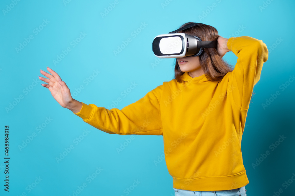 VR gamer, a young woman in a youth outfit uses a virtual reality helmet. Brunette in a yellow hoodie on a blue background with a virtual reality helmet on her head, full immersion VR gamer, a young woman in a youth outfit uses a virtual reality helmet. Brunette in a yellow hoodie on a blue background with a virtual reality helmet on her head, full immersion