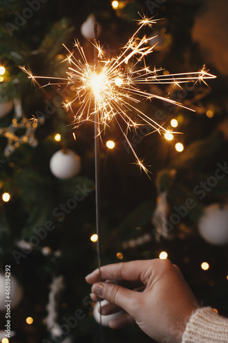 Happy New Year! Hand holding burning sparkler on background of christmas tree lights in eve