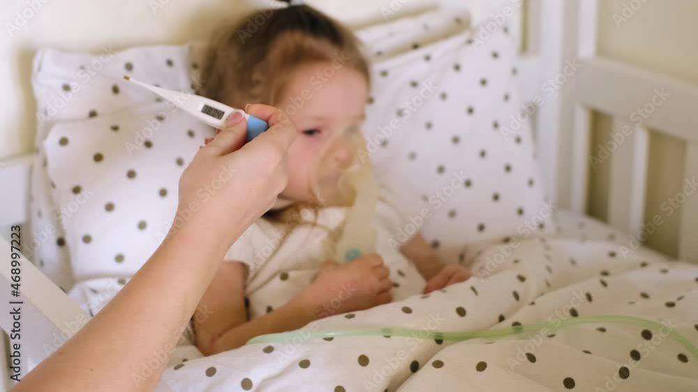 Mother takes out a thermometer with a high temperature from the armpit of a sick child. The girl makes inhalations, inhaling vapors through a mask with a tube, lying in bed. Seasonal viral diseases Mother takes out a thermometer with a high temperature from the armpit of a sick child. The girl makes inhalations, inhaling vapors through a mask with a tube, lying in bed. Seasonal viral diseases
