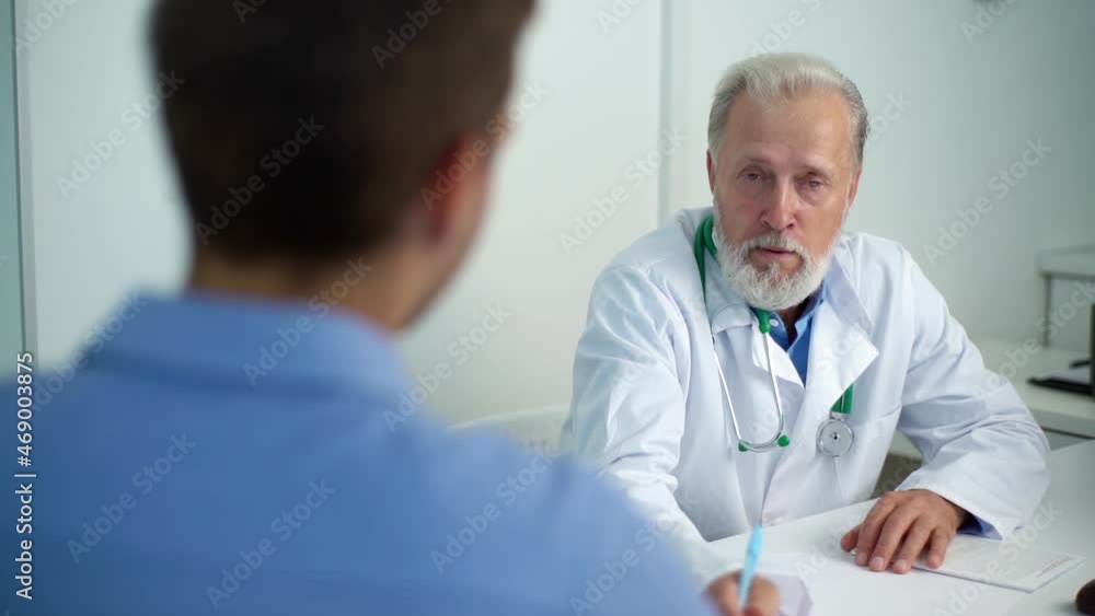 Close-up view from back to mature adult male doctor in medical coat consulting unrecognizable male patient about illness or surgery sitting at table, in medical office with light interior, slow motion Close-up view from back to mature adult male doctor in medical coat consulting unrecognizable male patient about illness or surgery sitting at table, in medical office with light interior, slow motion