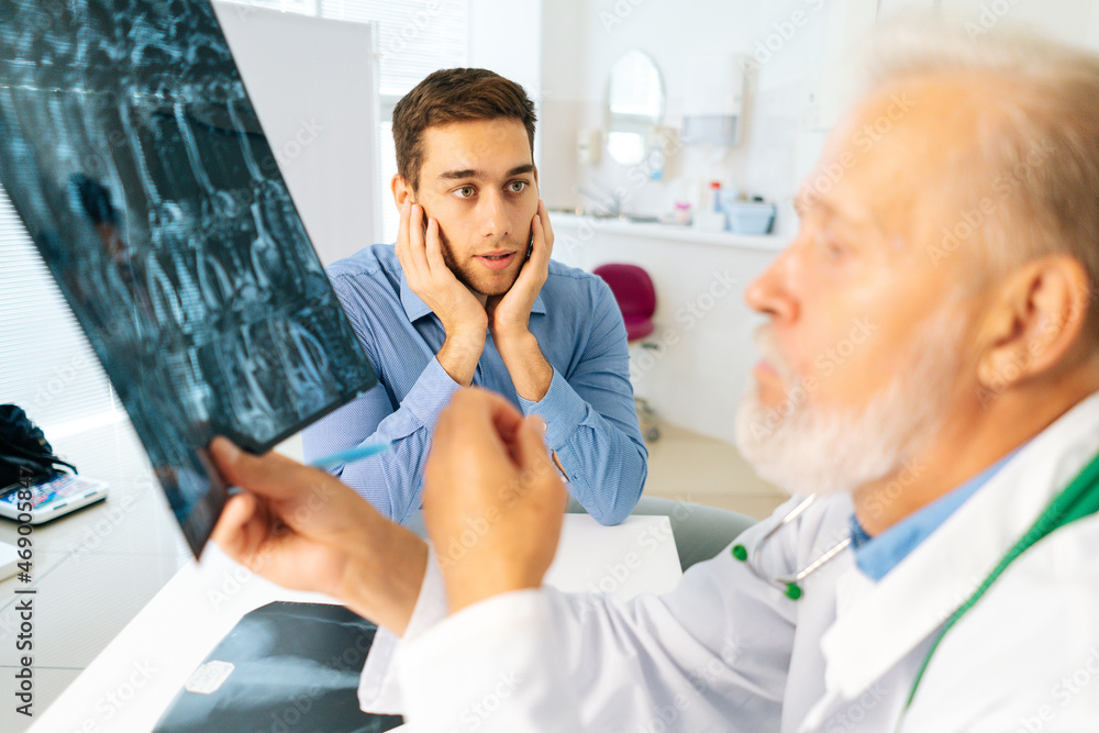 Obraz premium Close-up of mature adult male physician consult scared young man patient giving bad news explaining results of MRI image. Unhappy frustrated young man listening to bad news sitting in doctor office.
