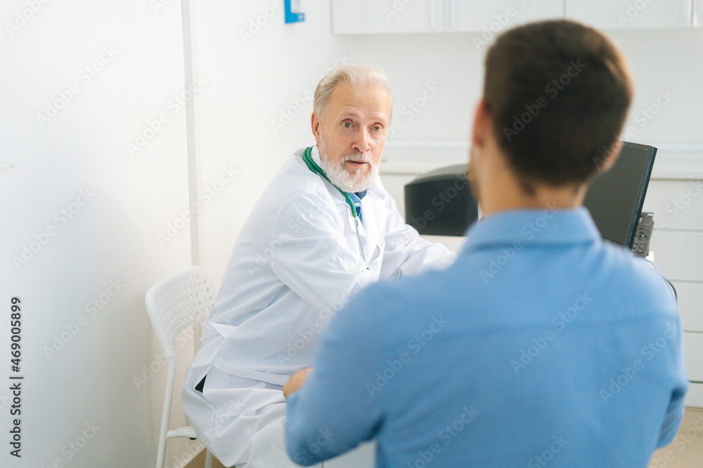 Close-up view from back to mature adult male doctor wearing white coat consulting unrecognizable young man patient about illness or surgery sitting at table, in medical office with light interior. Close-up view from back to mature adult male doctor wearing white coat consulting unrecognizable young man patient about illness or surgery sitting at table, in medical office with light interior.