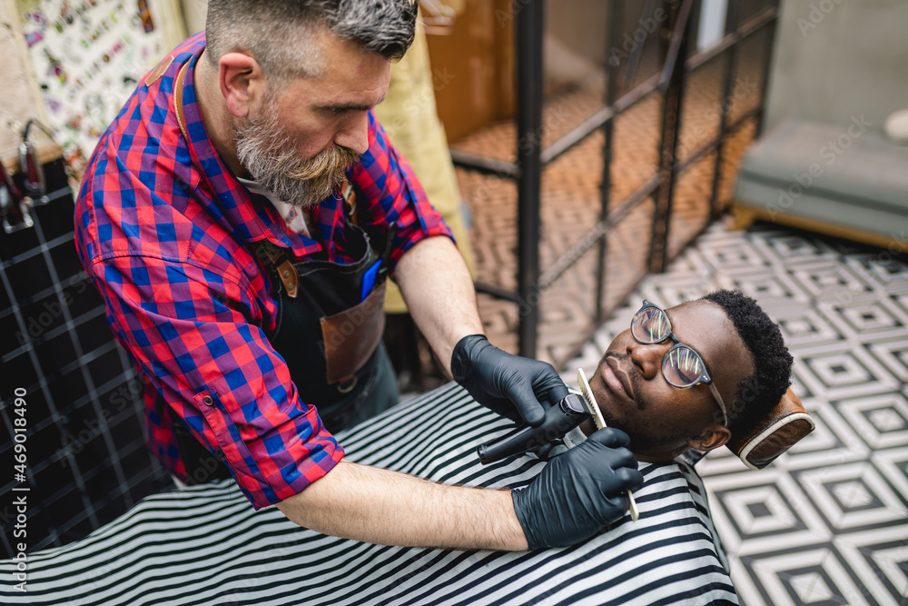 Obraz premium Young African man visiting hairstylist in barber shop. Professional hairdresser shaving beard with electric shearer machine.