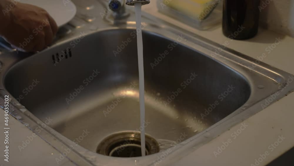 Person turns on the water and washes a white plate with a sponge in an iron sink in kitchen. Close-up. Man squeezes detergent onto plate and washes it in rays of warm artificial light. slow motion. Person turns on the water and washes a white plate with a sponge in an iron sink in kitchen. Close-up. Man squeezes detergent onto plate and washes it in rays of warm artificial light. slow motion.