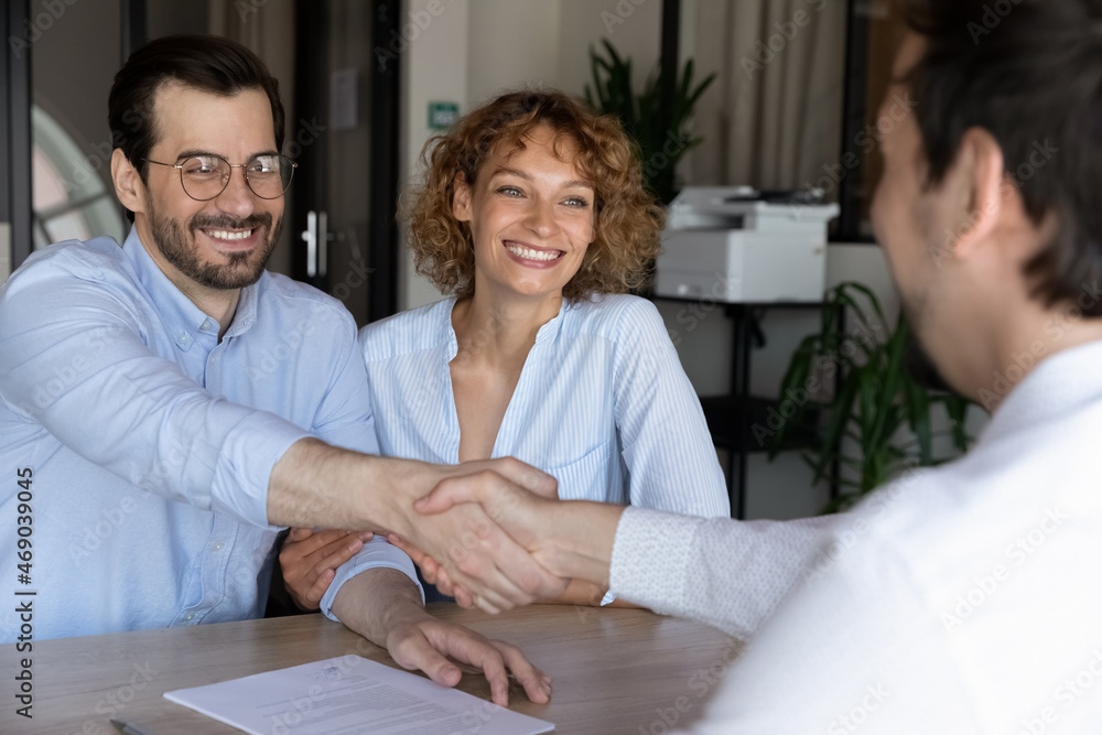 Obraz premium Joyful beautiful young family couple shaking hands with hispanic lawyer or broker, feeling thankful for professional consultation. Happy clients making agreement with realtor at office meeting.