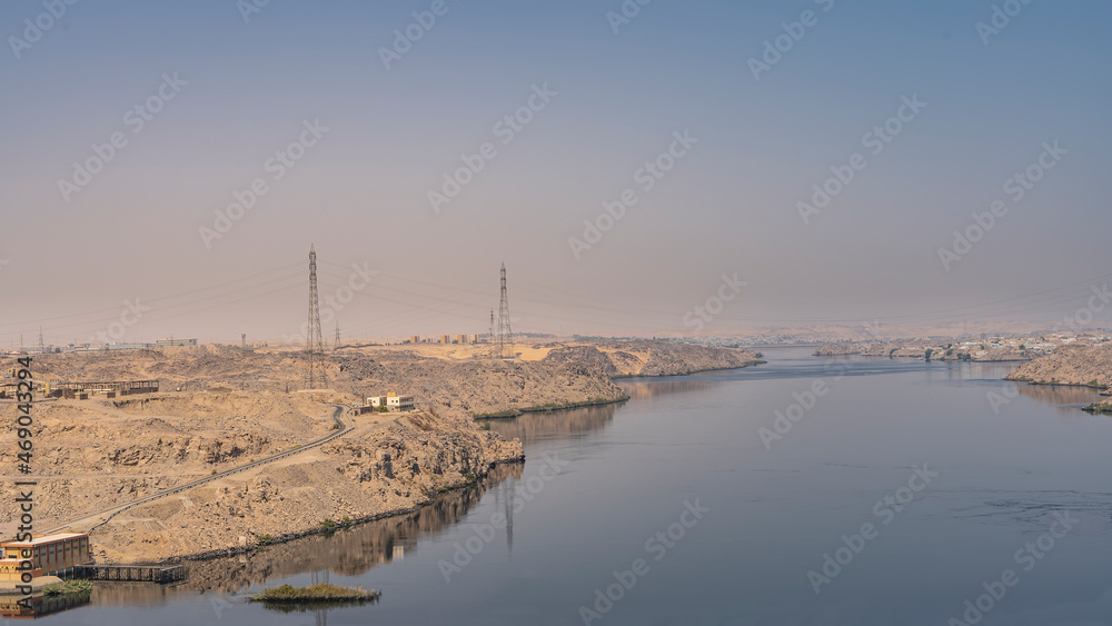The Nile River in the area of the Aswan Dam. Technical buildings are visible on the sandy shores. Blue sky. Reflection on the smooth surface of the water. Egypt