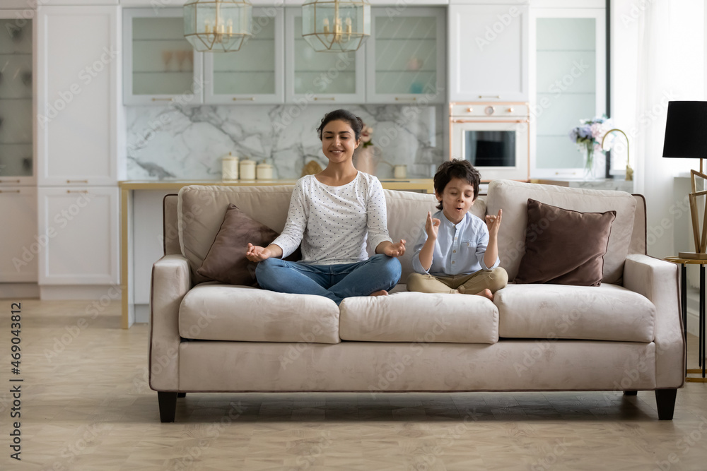 Happy calm Indian mom and little son kid sitting on couch, keeping lotus pose, zen hand gesture, meditating, practicing home yoga, home yoga, mental control, mindfulness, relaxing, exercising
