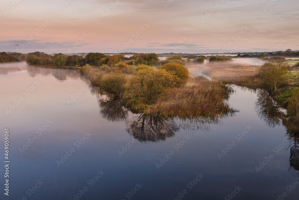 Nature scene. Calm water and trees reflection. Fog over meadow in the background. Calm and relaxing atmosphere. Soft pastel cloudy sky. View on a River Corrib, Galway city, Ireland. Nature scene. Calm water and trees reflection. Fog over meadow in the background. Calm and relaxing atmosphere. Soft pastel cloudy sky. View on a River Corrib, Galway city, Ireland.