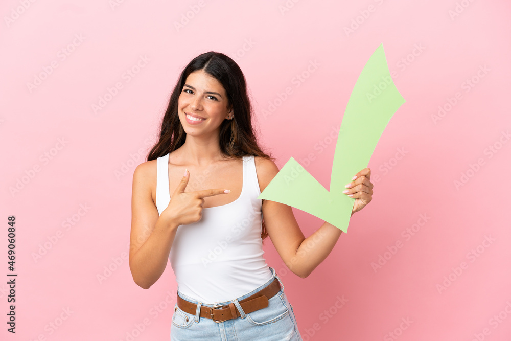 Young caucasian woman isolated on pink background holding a check icon and pointing it Young caucasian woman isolated on pink background holding a check icon and pointing it