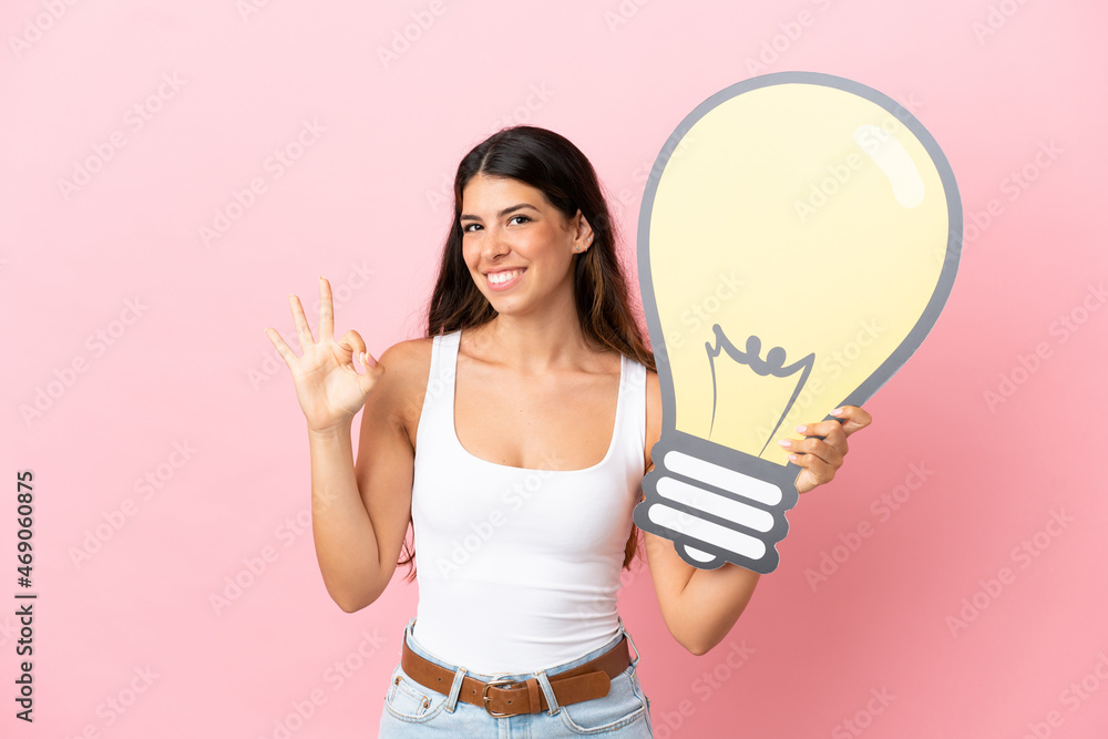 Young caucasian woman isolated on pink background holding a bulb icon and doing OK sign Young caucasian woman isolated on pink background holding a bulb icon and doing OK sign