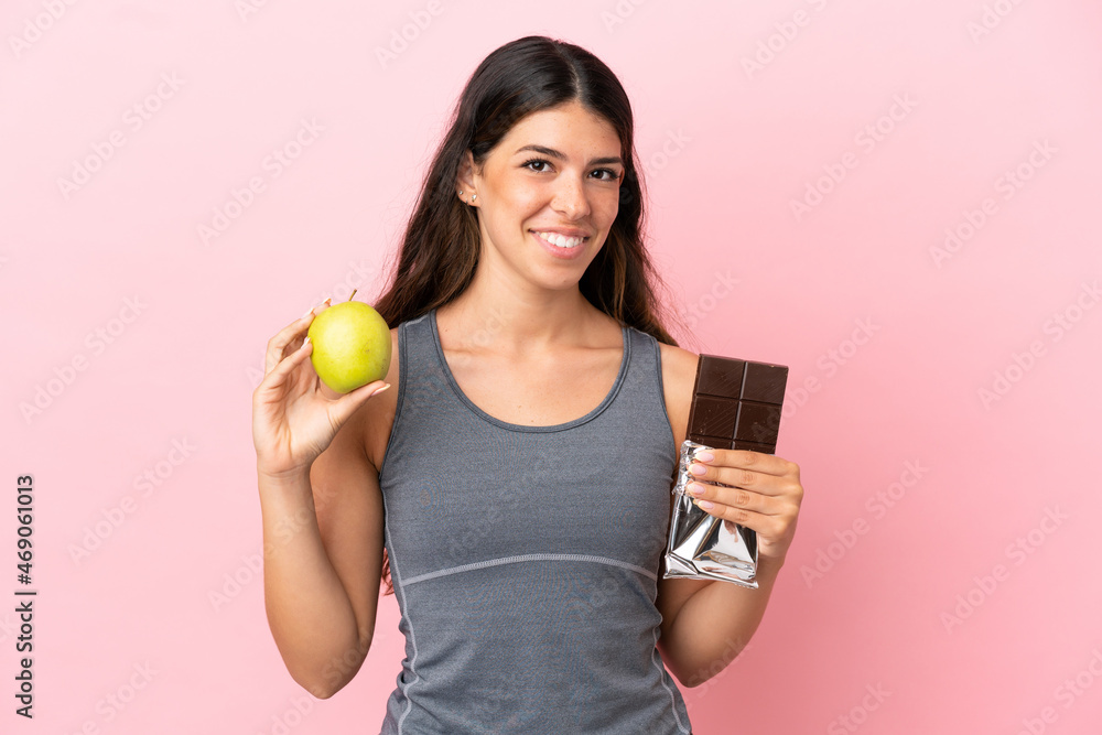 Young caucasian woman isolated on pink background taking a chocolate tablet in one hand and an apple in the other Young caucasian woman isolated on pink background taking a chocolate tablet in one hand and an apple in the other