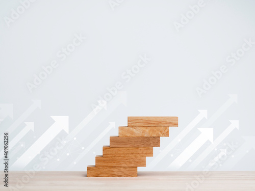 White rise up arrows with wooden blocks arranged as a steps chart on a wooden desk and clean white background with copy space  minimal style. Business growth process and economic improvement concept.