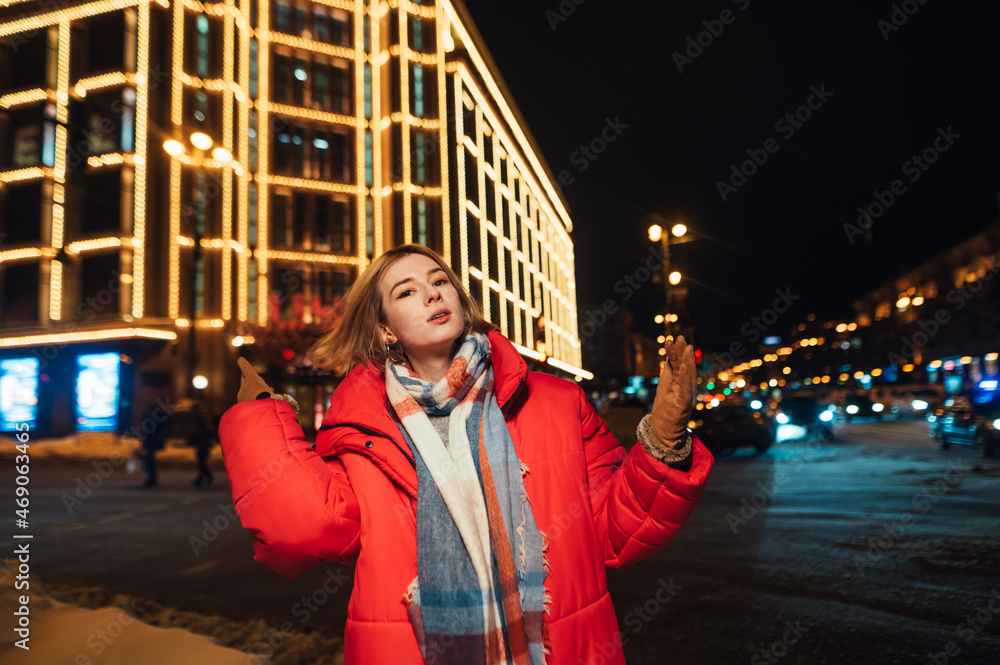 Fototapeta premium Attractive lady in a red warm jacket stands on the street against the backdrop of the cityscape and decorated with lanterns architecture for Christmas.