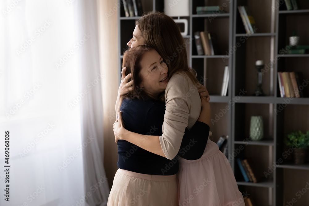 Happy mature woman mother with grown-up daughter hugging enjoying tender moment together, loving caring senior grandmother and granddaughter cuddling, standing in modern living room at home