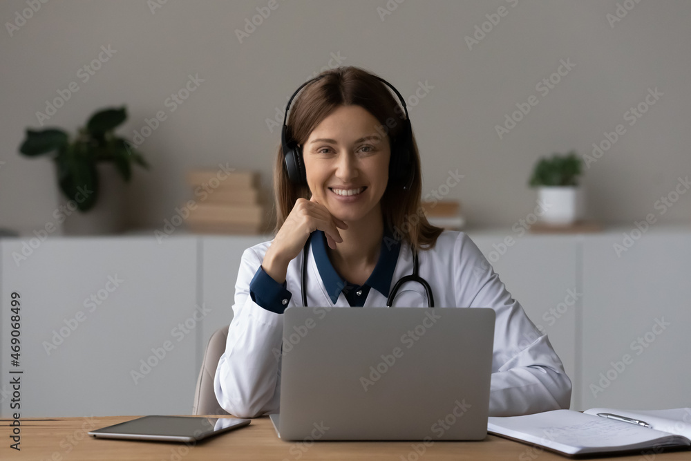 Head shot portrait smiling female doctor therapist in wireless headphones sitting at work desk with laptop, professional practitioner gp looking at camera, consulting patient online by video call Head shot portrait smiling female doctor therapist in wireless headphones sitting at work desk with laptop, professional practitioner gp looking at camera, consulting patient online by video call