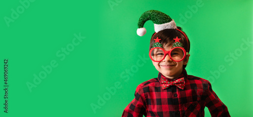 Little smiling boy in Christmas headband of Santa, funny Christmas glasses and Christmas bow-tie against green background