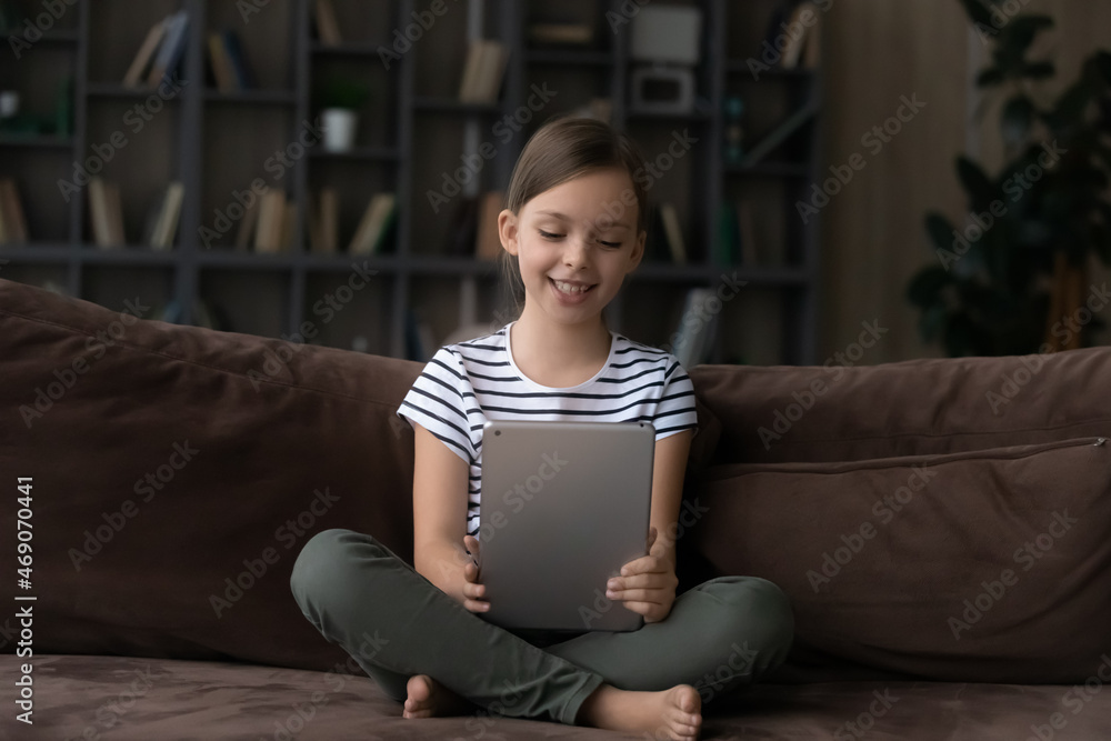 Happy cute girl kid using computer tablet, sitting on couch at home, smiling adorable child looking at device screen, having fun online, watching cartoons, video in social network, playing game Happy cute girl kid using computer tablet, sitting on couch at home, smiling adorable child looking at device screen, having fun online, watching cartoons, video in social network, playing game