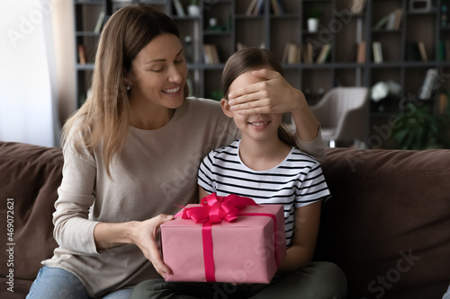 Loving mother covering little daughter eyes, congratulating with birthday, preparing surprise, adorable cute girl holding pink gift box, family celebrating special event, sitting on couch at home
