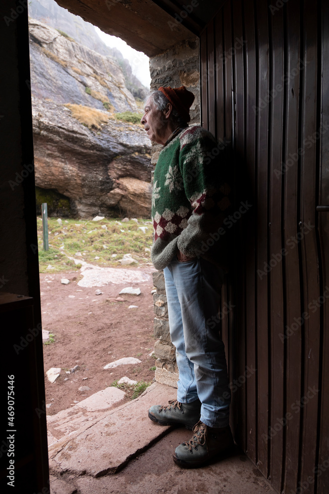 A mountaineer, senior adult man, in his 70s, with handmade woolen sweater, observes the outside from the door of a mountain hut, Pyrenees, Spain