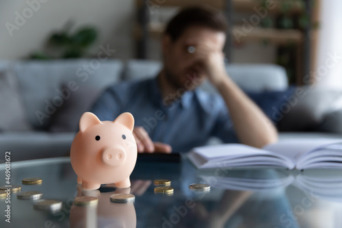 Close up focus on piggybank on table, young man feeling stressed calculating expenditures or taxes, managing future payments or planning investments, suffering from lack of money, accounting concept.