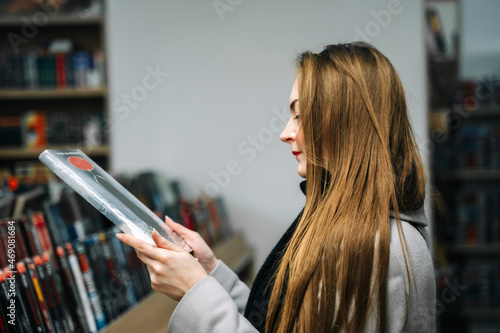 A beautiful young woman chooses a comic book for a gift in a bookstore to buy it. Stands in front of the bookshelf and looks at the comic book while holding it.