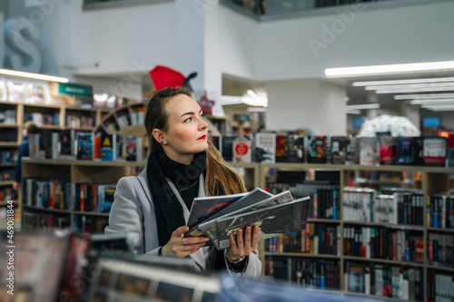 A beautiful young woman chooses a comic book for a gift in a bookstore to buy it. Stands in front of a shelf of books and holds a stack of comics comparing them.