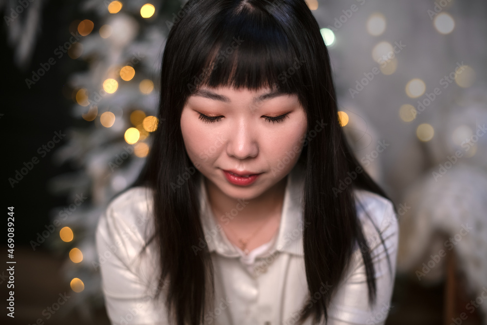 A young, beautiful Korean Asian woman with modestly lowered eyes is in her cozy home and dreaming against the background of a Christmas tree in the evening. The concept of New Year and Christmas A young, beautiful Korean Asian woman with modestly lowered eyes is in her cozy home and dreaming against the background of a Christmas tree in the evening. The concept of New Year and Christmas