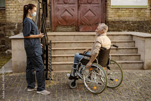 Full length shot of lovely young nurse in protective face mask helping mature disabled man in wheelchair outdoors